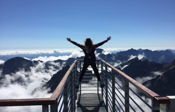 woman standing on the rail with arms spread looking down on mountains and clouds