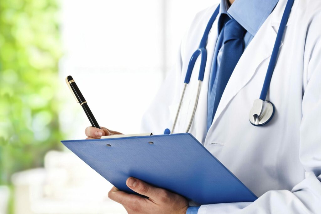 man in a lab coat wearing a blue stethoscope and writing on a blue clipboard